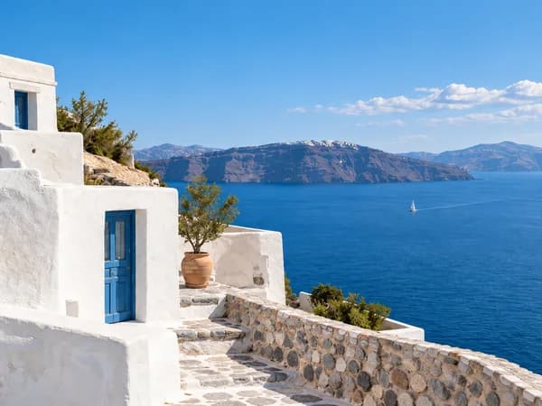 White coastal buildings with blue doors overlooking a blue bay and a sailboat